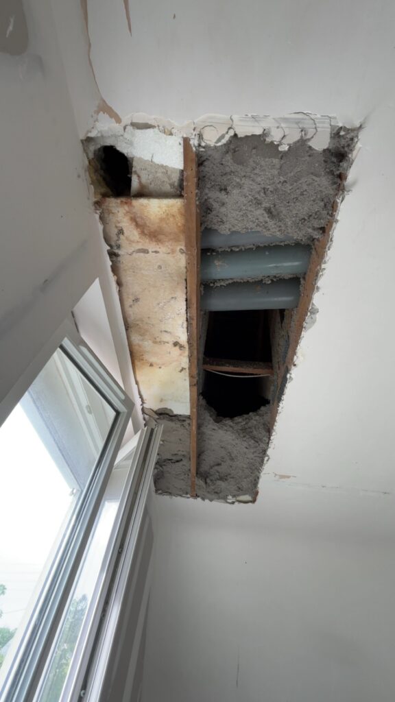 Réparation de Plafond : Isolation et Tuyaux Apparents Trou dans un plafond révélant de l'isolation grise, des solives en bois et des tuyaux en PVC près d'une fenêtre.