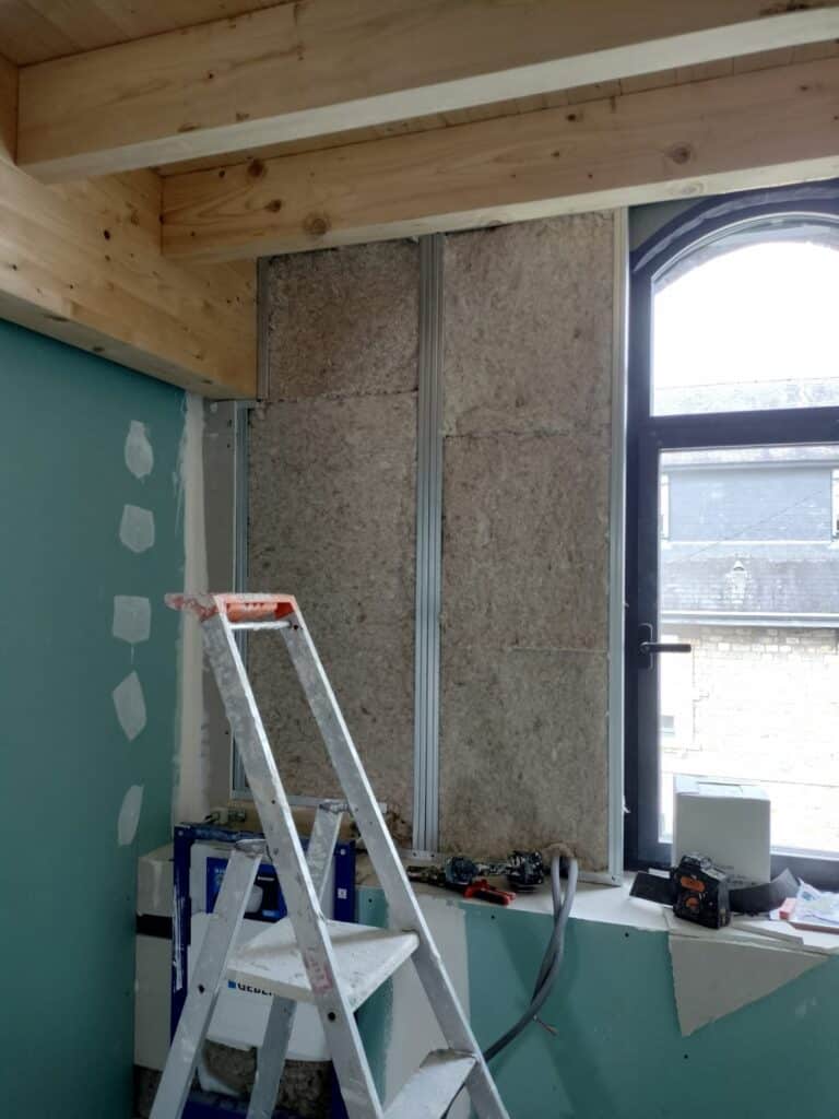 Isolation Murale et Placo : Chantier Intérieur en Cours Construction intérieure : isolation en laine minérale entre montants métalliques près d'une fenêtre arquée, avec un escabeau et des plaques de plâtre partiellement jointées.