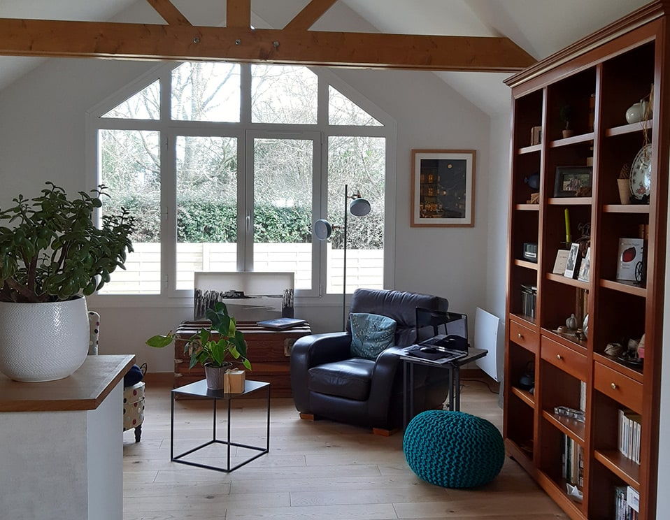 Salon cosy avec grande baie vitrée et bibliothèque Salon lumineux avec grande fenêtre à pignon, fauteuil en cuir marron, bibliothèque en bois et plantes vertes, décor cosy.