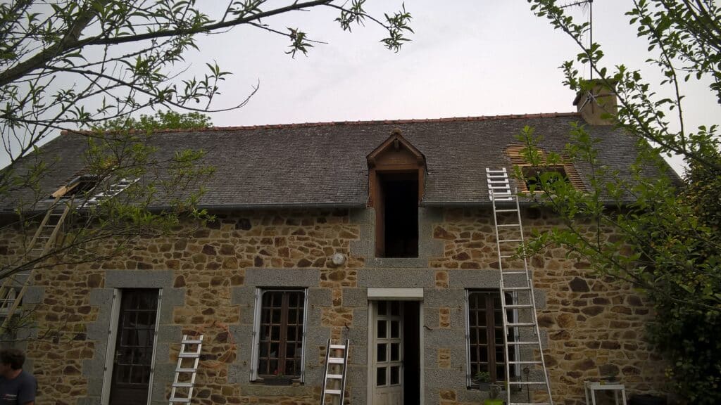 Ancienne façade en pierre de moellons et granit, toit en ardoise. Rénovation de toiture en cours avec échelles et lucarnes ouvertes.